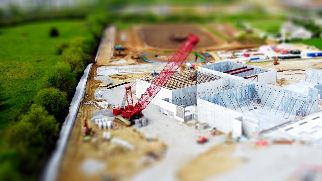 services-04 Aerial view of a construction site with a red crane amidst green fields and concrete buildings.